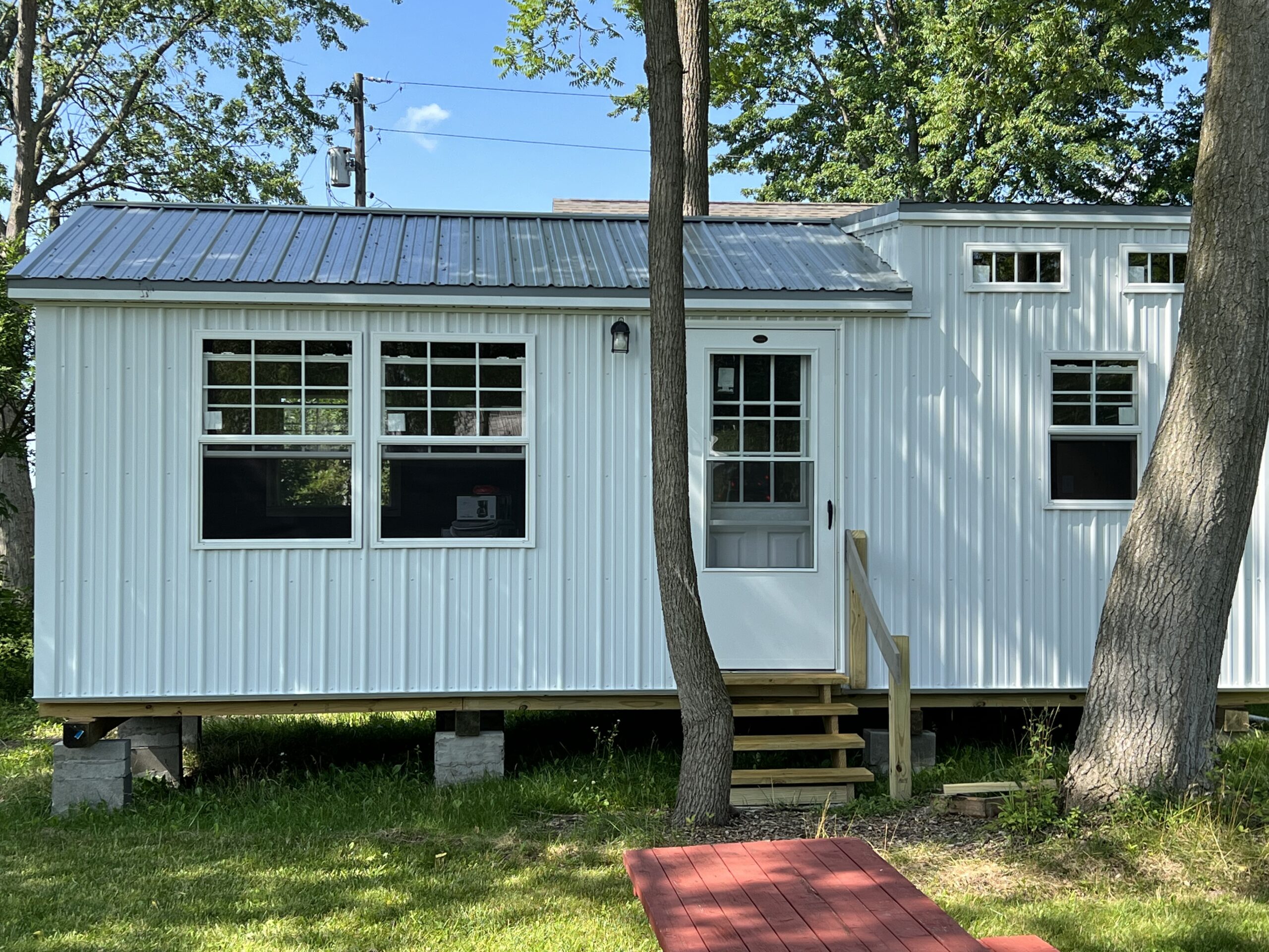 This image showcases a 12x18 modern cabin with a sleek white exterior and a metal roof, giving it a contemporary yet functional appearance. The cabin features large double windows on one side, allowing for ample natural light, and smaller accent windows for added ventilation and charm. A central door with a glass panel leads to a small wooden staircase, enhancing accessibility. The cabin is elevated on a sturdy foundation of concrete blocks, blending effortlessly with its lush green surroundings. Perfect for use as a vacation retreat, studio space, or tiny home.