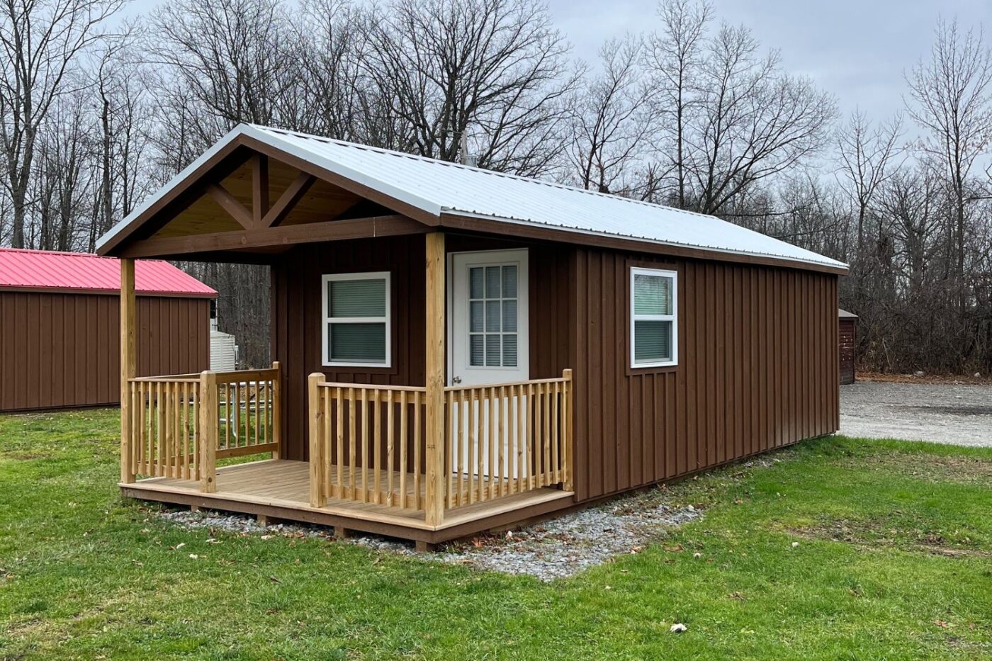 The image depicts a charming 14x20 gable porch cabin featuring a wooden exterior stained in a warm brown tone. The front includes a small porch with natural wood railings and posts, complementing the rustic aesthetic. A white door with a glass panel and a side window provide light and ventilation. This cozy cabin sits on a gravel foundation surrounded by a grassy area, blending seamlessly into the serene countryside backdrop. Ideal for various uses, such as a guesthouse, home office, or a relaxing retreat. The image depicts a charming 12x18 gable porch cabin featuring a wooden exterior stained in a warm brown tone. The front includes a small porch with natural wood railings and posts, complementing the rustic aesthetic. A white door with a glass panel and a side window provide light and ventilation. This cozy cabin sits on a gravel foundation surrounded by a grassy area, blending seamlessly into the serene countryside backdrop. Ideal for various uses, such as a guesthouse, home office, or a relaxing retreat.