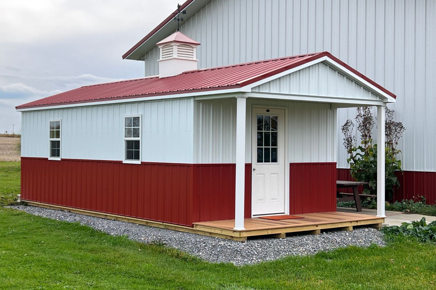 A charming gable porch cabin with a red and white metal exterior, featuring a bold red roof and white trim accents. The structure includes a small covered porch with sturdy white columns and a wooden deck, creating a welcoming entrance. The cabin is equipped with tall white-framed windows and a white entry door with glass panels, adding a touch of elegance. Positioned on a gravel foundation and surrounded by green grass, the cabin sits near a larger matching structure, making it a functional and aesthetically pleasing addition to the landscape. A decorative cupola on the roof adds a classic barn-inspired detail.