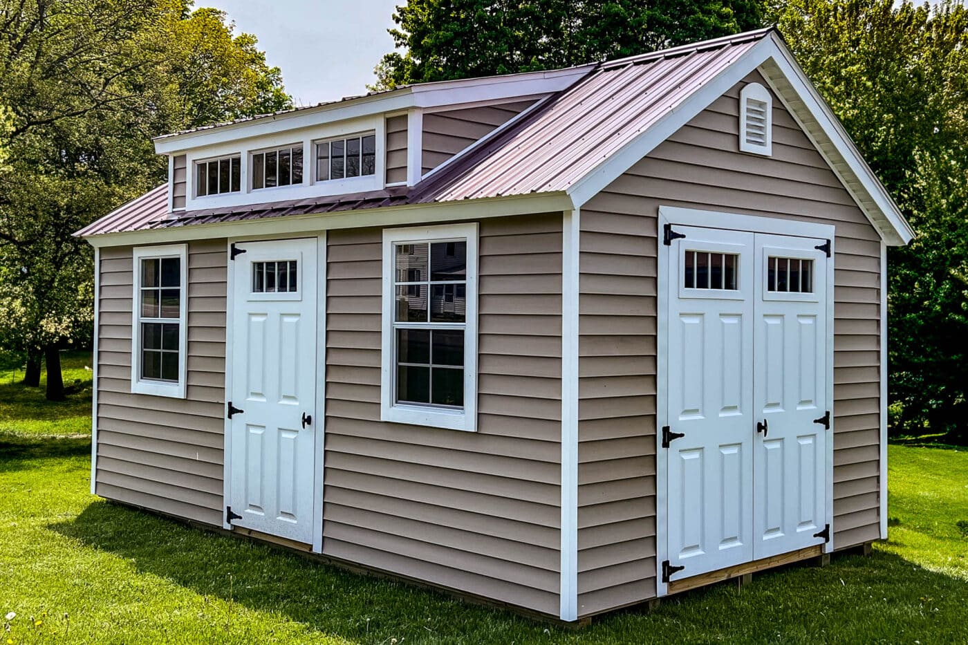 Tan vinyl-sided shed with a red metal roof, two white single-entry doors, and a set of white double barn-style doors. The shed includes a row of transom windows and is situated on a grassy lawn with trees in the background.