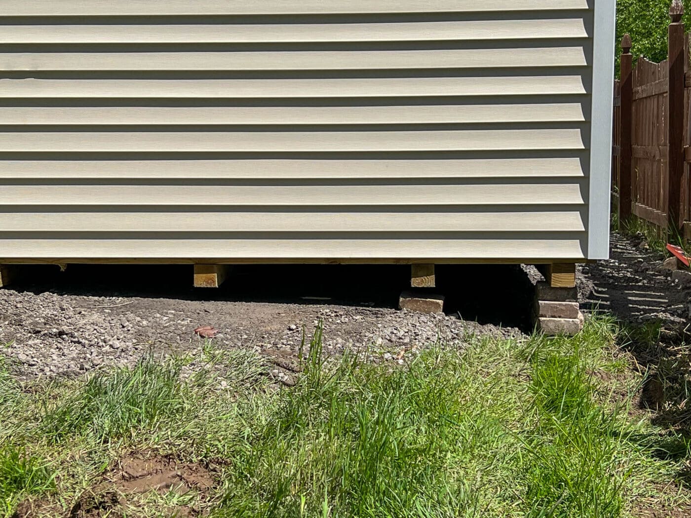 A side view of a beige shed elevated on wooden blocks with a gravel base underneath. The shed's vinyl siding and the gravel foundation are visible.