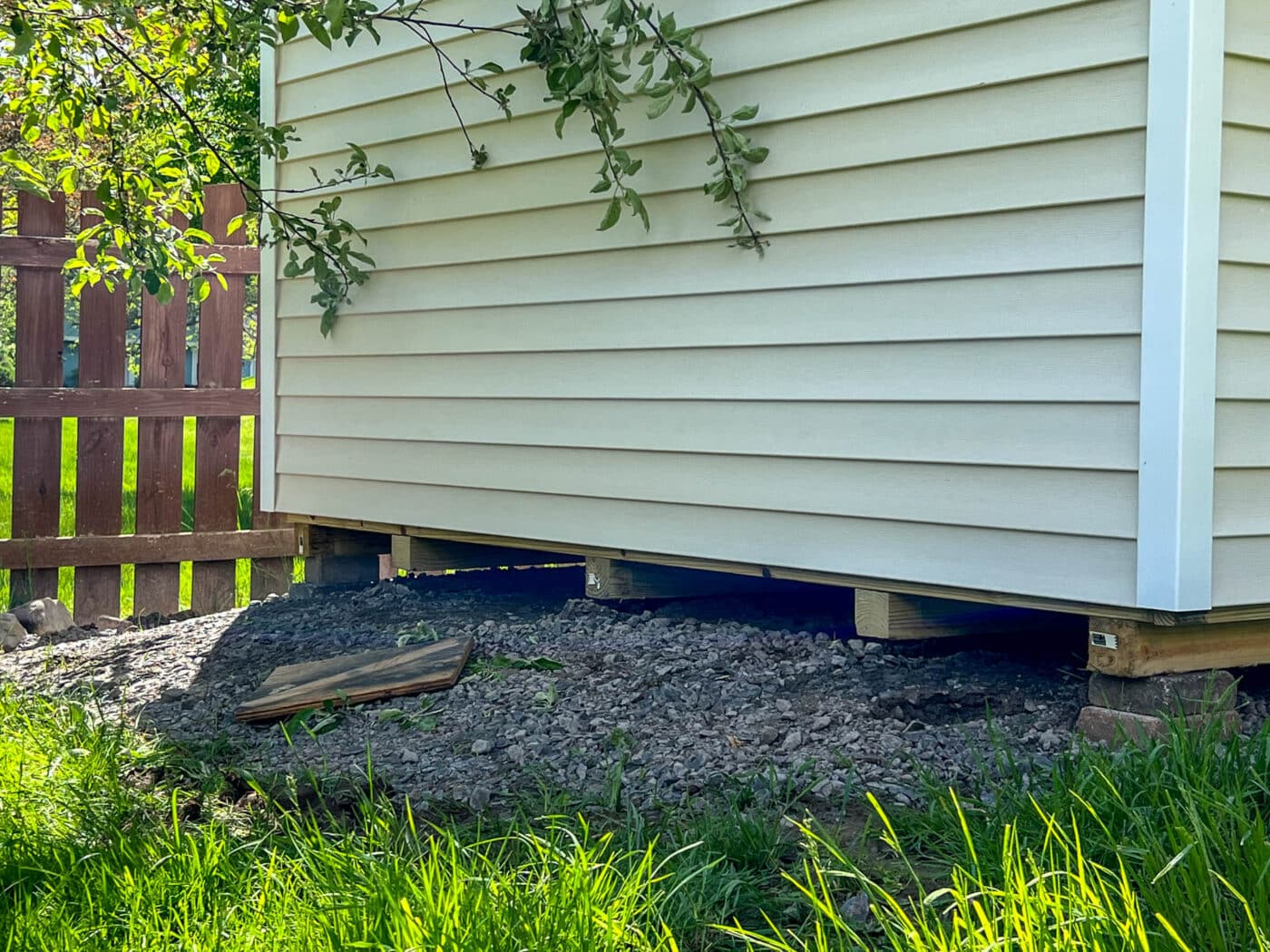 A close-up of a beige vinyl-sided shed elevated on wooden blocks, showing the gravel foundation underneath. The shed is positioned near a wooden fence with grass and small plants surrounding the base.