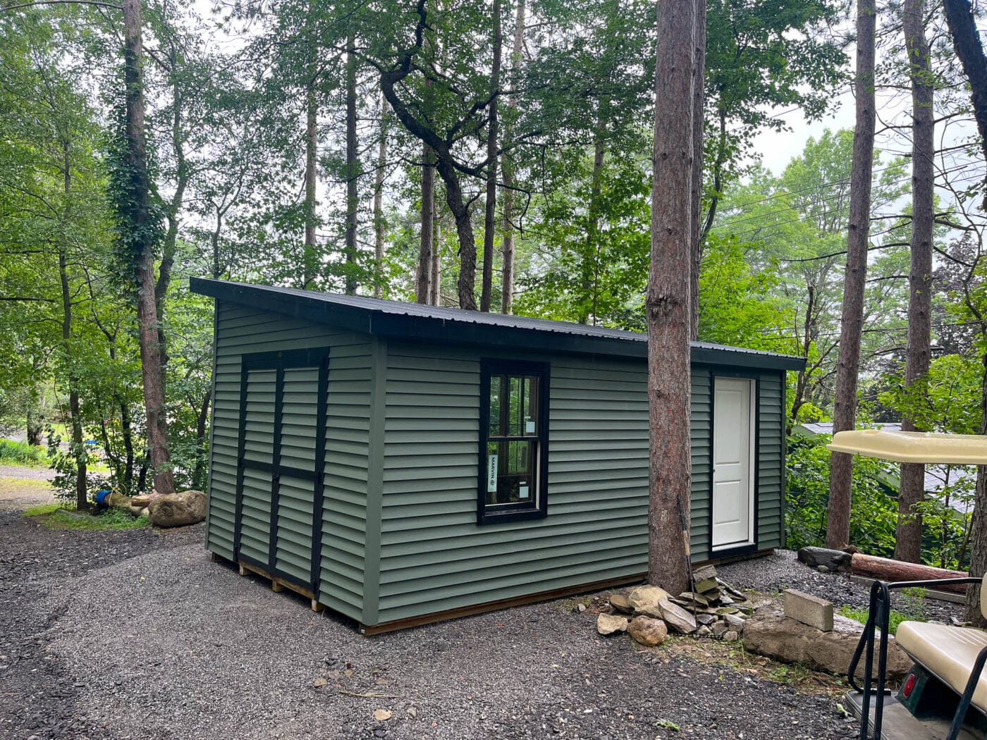 A green shed with black trim and a black metal roof, elevated on a well-leveled gravel base surrounded by trees. A white door and black-framed window are visible on the side.