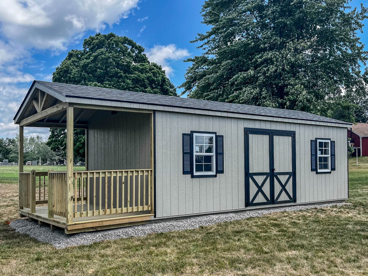 A beige shed with a covered porch supported by wooden posts, a gray gable roof, and double barn-style doors with black hinges and white trim. The shed is surrounded by grass and trees under a clear sky.