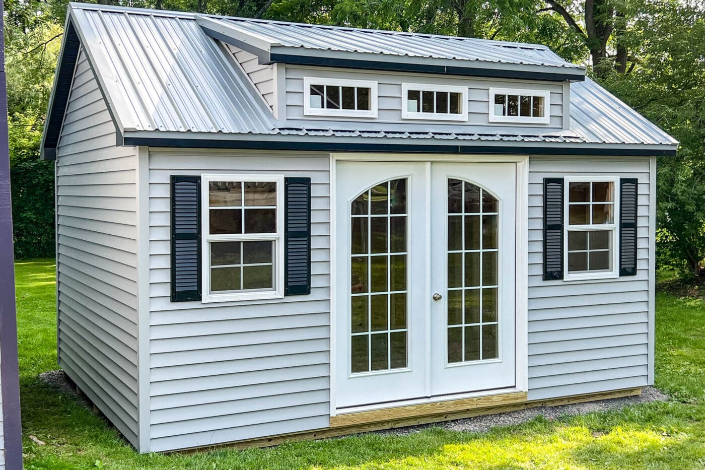 A small, gray-sided structure by Sheds By Fisher featuring French doors, transom windows, black shutters, and a metal roof, set on a grassy lot with trees in the background.
