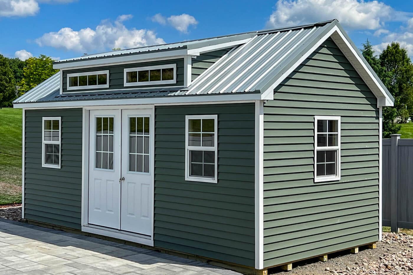 Green vinyl-sided shed with white double doors, transom windows, and a metal roof, built by Sheds By Fisher. The structure sits on a gravel pad and is surrounded by grass and trees under a bright blue sky.