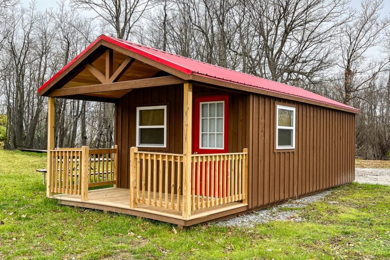 Rustic brown portable cabin shed with a red metal roof and front porch, set on grass near trees.