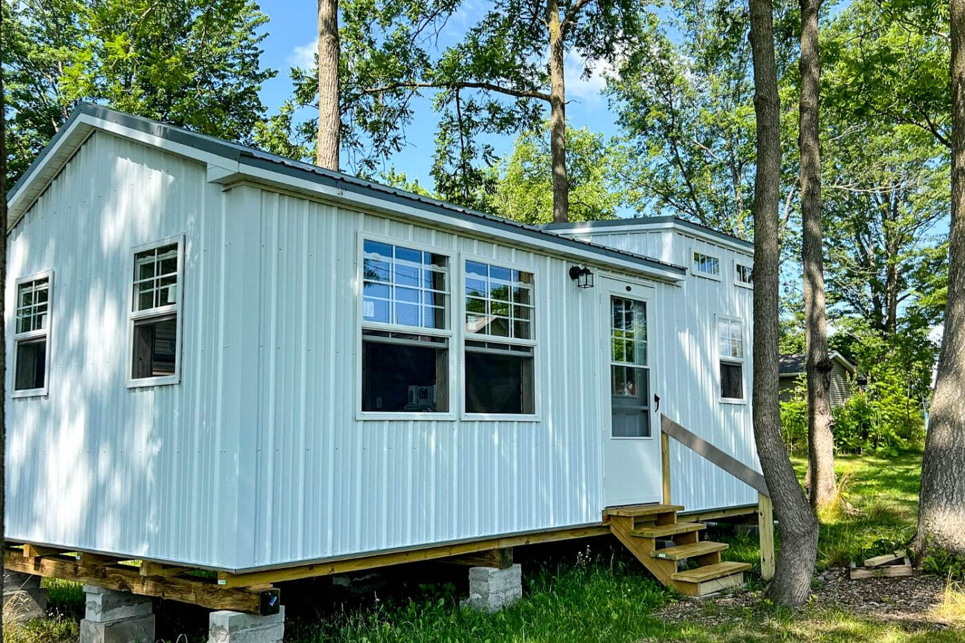 "White portable shed with black trim and a small porch, set among green trees."