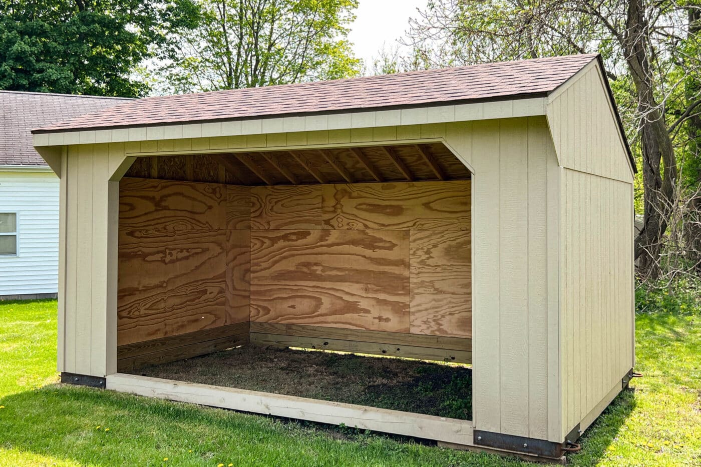 Beige run-in shed with an open front, wooden interior, and shingled roof on a green lawn.