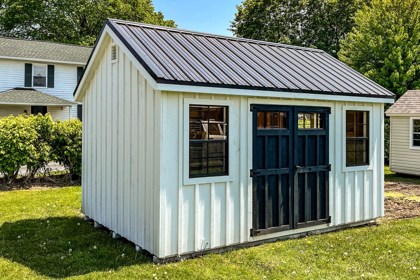 White chalet-style storage shed with black doors and windows near a residential home. Just one of our many sheds near East Aurora NY.