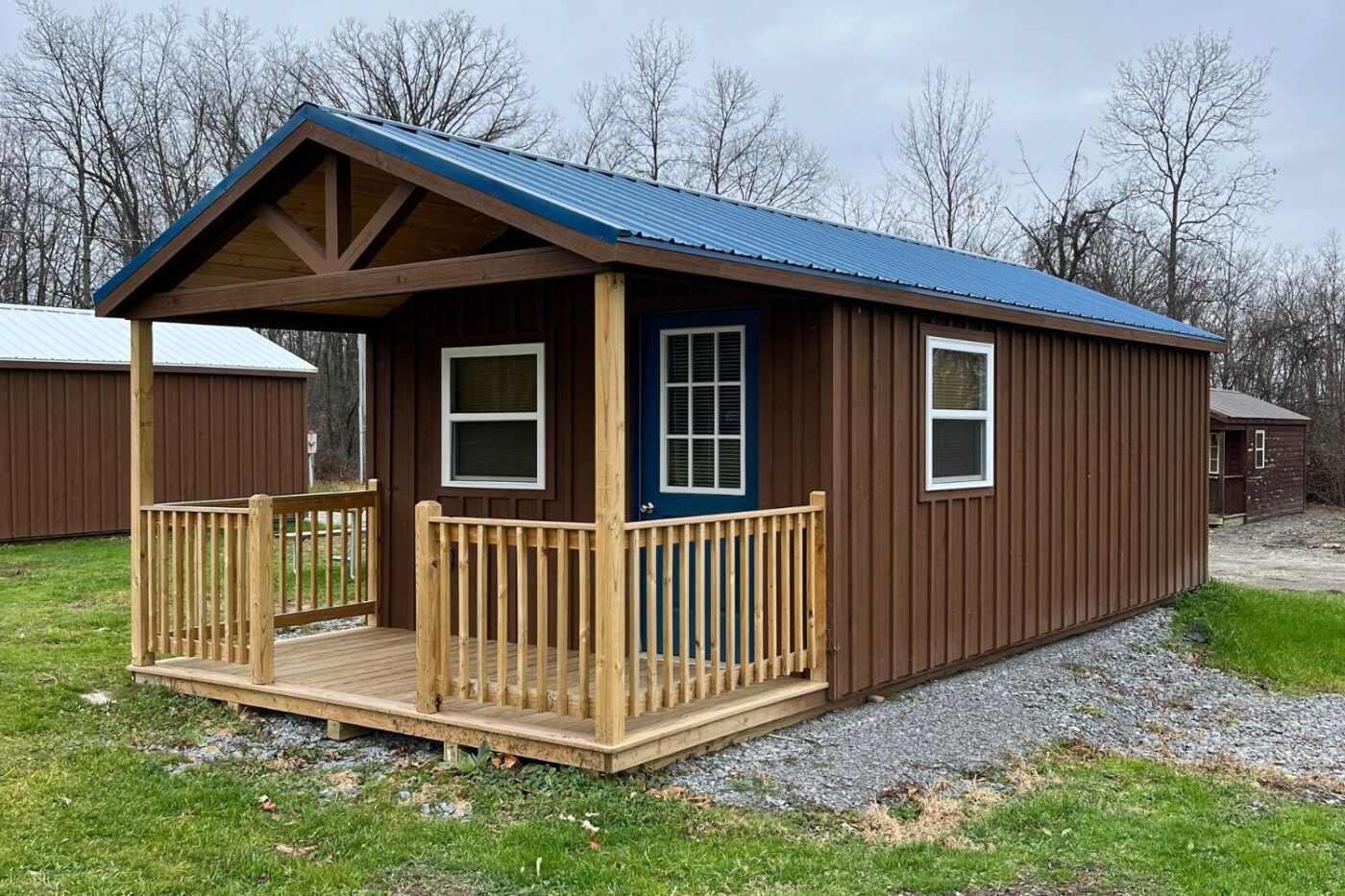 The image depicts a charming 14x24 gable porch cabin featuring a wooden exterior stained in a warm brown tone. The front includes a small porch with natural wood railings and posts, complementing the rustic aesthetic. A white door with a glass panel and a side window provide light and ventilation. This cozy cabin sits on a gravel foundation surrounded by a grassy area, blending seamlessly into the serene countryside backdrop. Ideal for various uses, such as a guesthouse, home office, or a relaxing retreat. The image depicts a charming 14x24 gable porch cabin featuring a wooden exterior stained in a warm brown tone. The front includes a small porch with natural wood railings and posts, complementing the rustic aesthetic. A white door with a glass panel and a side window provide light and ventilation. This cozy cabin sits on a gravel foundation surrounded by a grassy area, blending seamlessly into the serene countryside backdrop. Ideal for various uses, such as a guesthouse, home office, or a relaxing retreat.