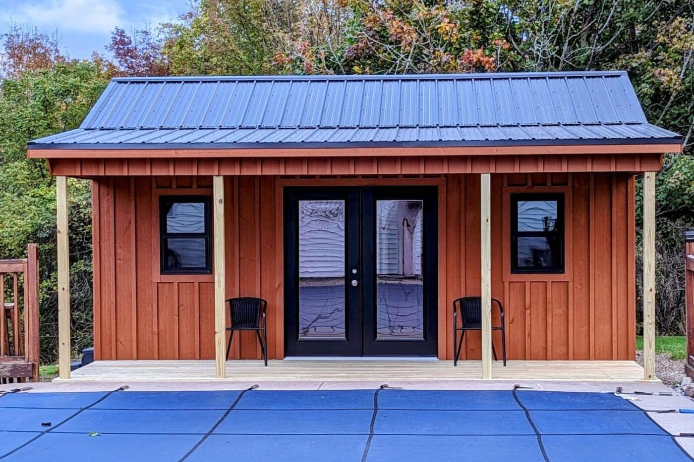 A stylish 14x20 cabin featuring rich, red-stained wood siding and a sleek black metal roof. The structure includes a charming covered porch supported by natural wooden posts, providing a welcoming outdoor space. Two black-framed windows flank elegant black double French doors, creating a balanced and modern design. The porch is furnished with a pair of black chairs, perfect for relaxation. Positioned adjacent to a covered pool and surrounded by vibrant autumn foliage, the cabin offers a cozy and functional retreat with a touch of contemporary flair.